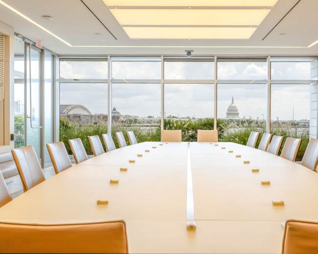 A large, modern conference room features a long table surrounded by chairs, with large windows showcasing a view of an urban landscape and a dome structure in the background.