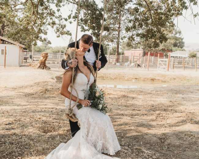A bride in a lace dress sits on a swing while being embraced by a groom standing behind her in a suit.