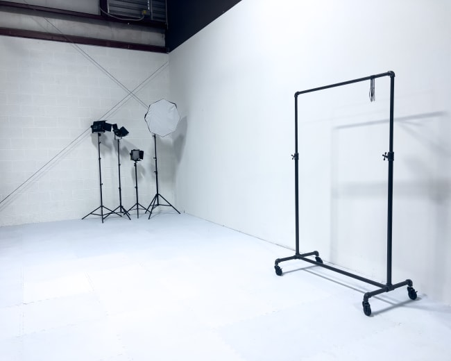 The image shows a studio space with a clothing rack on wheels and three lighting setups, including a softbox, against a blank wall.