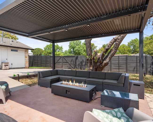 The image shows a modern outdoor patio area with a sectional seating arrangement, a fire pit, and a pergola overhead.
