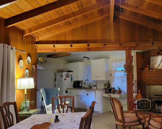 The image shows a rustic interior of a living space featuring a dining area with a white table, a kitchen area in the background, and a brick fireplace surrounded by wooden furnishings.
