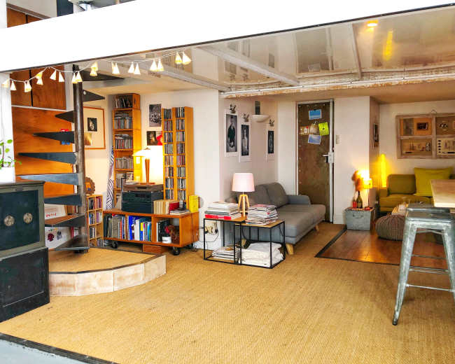 The image shows a modern living space featuring a sofa, bookshelves, a coffee table, and a warm light fixture, with a mix of textures and colors throughout the room.