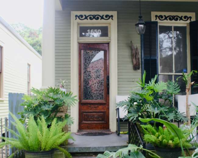 The image shows a front porch with a wooden door and steps flanked by large potted plants.