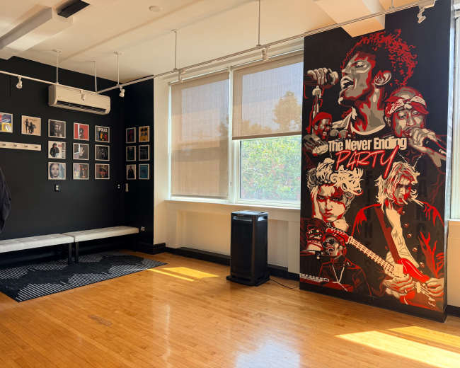 The room features a black wall adorned with framed art and a large, colorful mural of musicians, alongside a wooden floor and windows with shades.