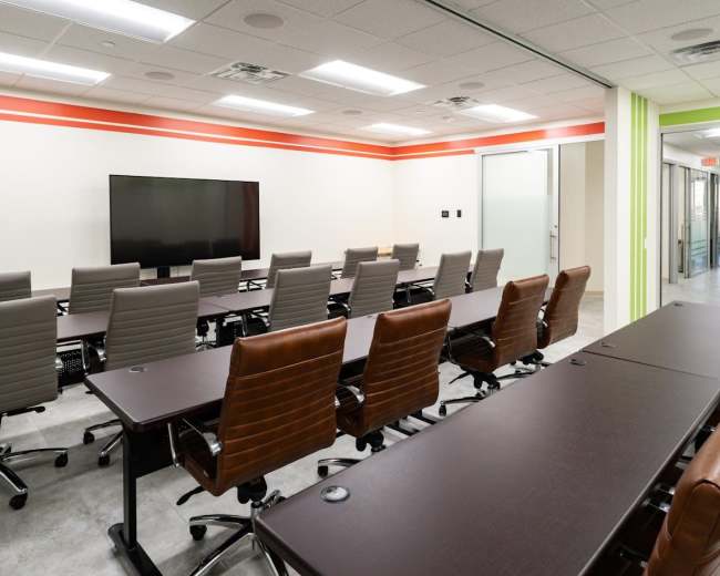 The image shows a modern conference room with several brown and gray chairs arranged around dark wooden tables, a large television screen on the wall, and glass doors leading to a corridor.