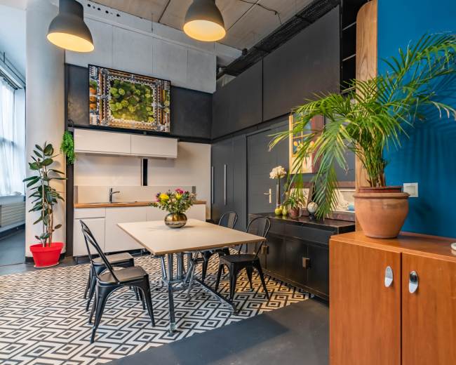 The image shows a modern kitchen and dining area featuring a table with chairs, a wall-mounted artwork, and a potted plant beside a wooden cabinet.