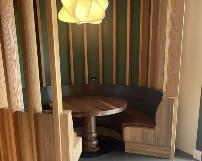The image shows a cozy circular seating booth with a wooden table surrounded by wooden slats and a pendant light overhead.