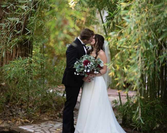 A couple is embracing and kissing under a canopy of lush green foliage, with one partner holding a bouquet.