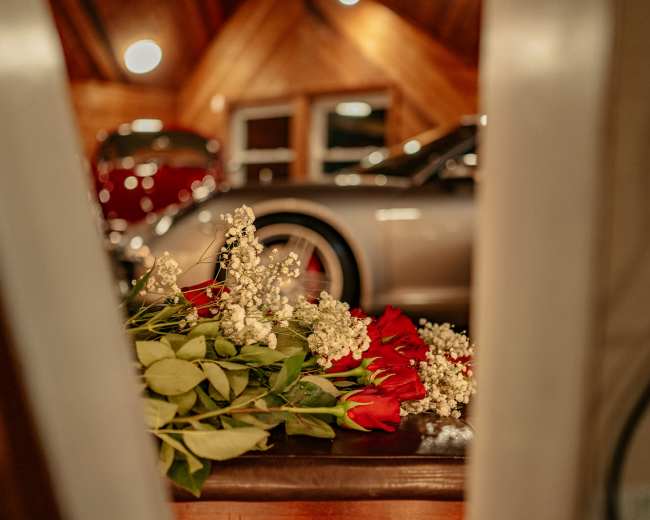 A bouquet of red roses and white flowers rests on a table near vintage cars parked in a wooden-paneled garage.