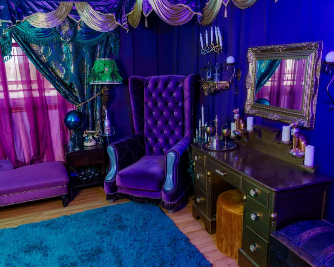 The image shows a richly decorated room featuring a purple velvet armchair, a matching chaise lounge, a vanity with a mirror, and curtains of deep blue and magenta hues.