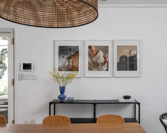 A dining area features a wooden table, wicker chairs, and a wall adorned with three framed artworks above a black console table, complemented by a blue vase with yellow flowers.