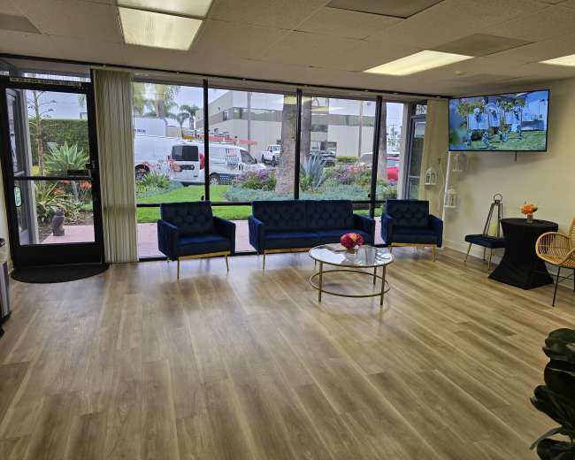 The image shows a modern waiting area with blue sofas, a round coffee table, a TV mounted on the wall, and large windows overlooking an outdoor space.