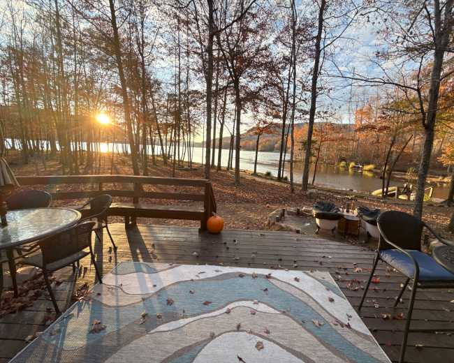 A wooden deck overlooking a lake is framed by trees with autumn foliage as the sun sets in the background.