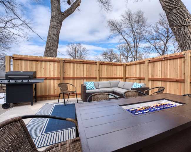 A cozy outdoor patio area with a fire pit, seating arrangement, and a barbecue grill surrounded by wooden fences and trees.