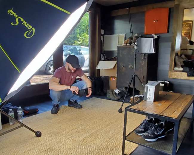 A person is crouching on a rug in a room with a large softbox light and a wooden table displaying shoes, while boxes and a safe are visible in the background.