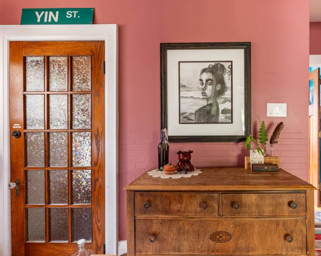The image depicts a hallway with a wooden dresser, a framed portrait on the wall, and a glass-paned door to the left.