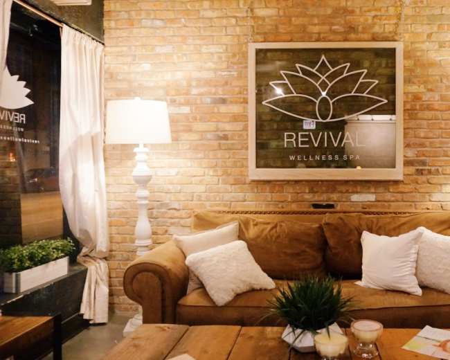 The image shows a cozy wellness spa interior with a brick wall, a framed sign featuring the word "REVIVAL," a brown sofa with white pillows, and two white lamps.