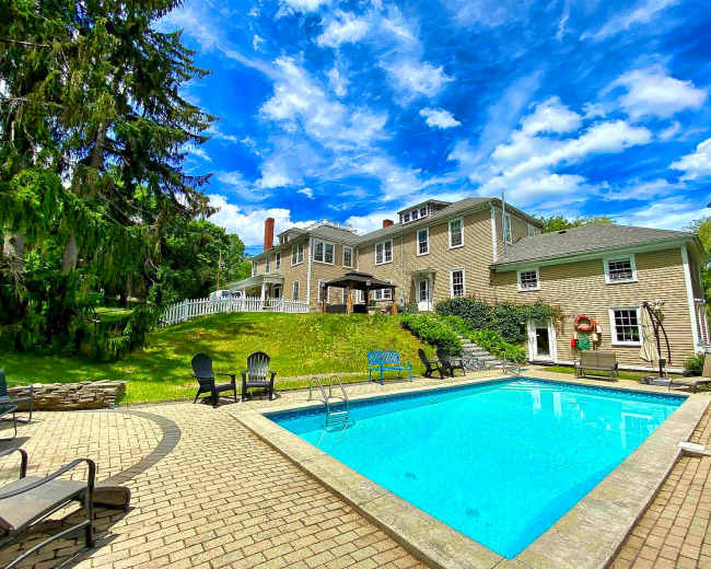 The image shows a swimming pool surrounded by a patio area and a large house set against a backdrop of blue sky and scattered clouds.