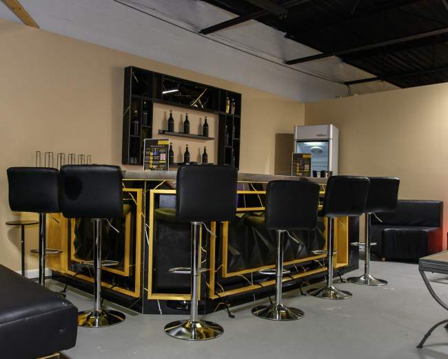 The image shows a modern bar area with a glossy black and gold countertop, high-backed stools, and a shelf displaying bottles in a minimalist setting.