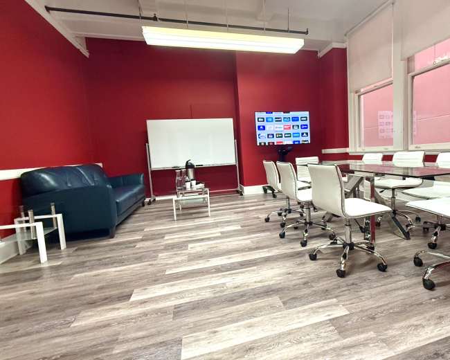 The image shows a meeting room with a red wall, a blue couch, a glass conference table surrounded by white chairs, a whiteboard, and a television screen mounted on the wall.