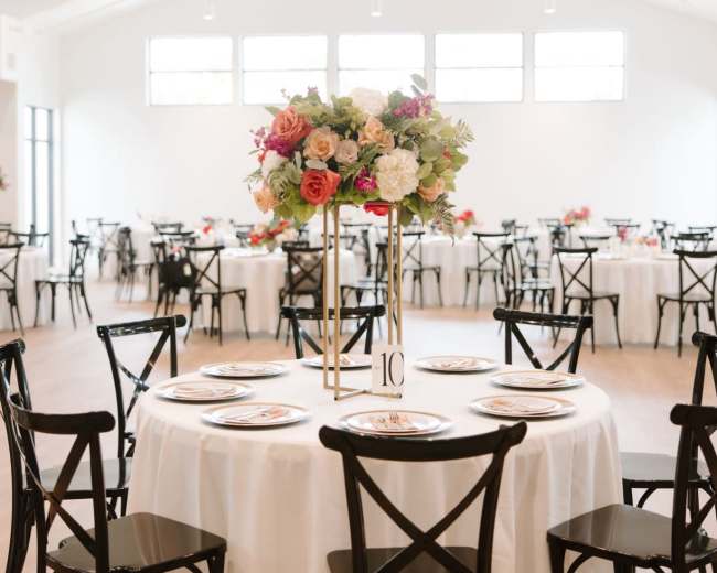 A spacious dining area features round tables with floral centerpieces and black wooden chairs, illuminated by natural light from large windows.