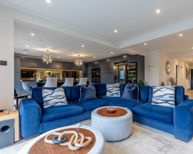 The image shows a modern living room with a large blue sectional sofa, a round coffee table, and a spacious kitchen in the background.