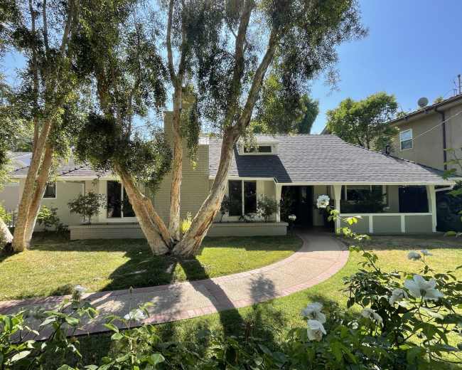A single-story house with a dark roof and large windows is set in a green yard with trees and a curved walkway.