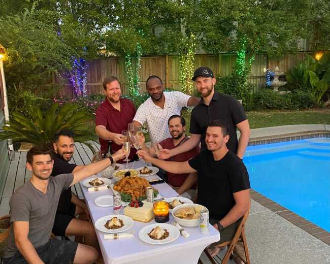 A group of seven men toast with drinks at a table set near a pool, surrounded by greenery and evening lights.