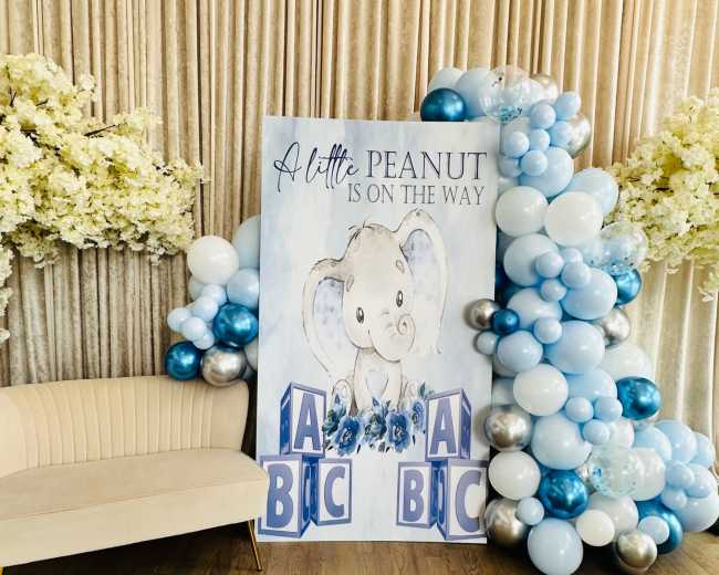 A decorative setup featuring a large sign with an elephant and the text "A little peanut is on the way," surrounded by clusters of blue and silver balloons and floral arrangements, with a cream-colored sofa in front.