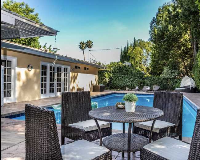 A circular outdoor table with four chairs is set beside a rectangular swimming pool surrounded by greenery and a yellow building in the background.