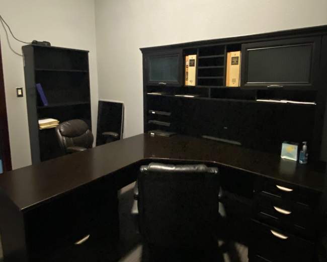 The image shows a dark wooden office setup with a large desk, an ergonomic chair, and a bookshelf in a dimly lit room.