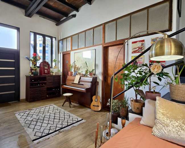 The image shows a cozy interior space featuring a piano, a guitar, plants, and a cozy seating area, with large windows providing natural light.