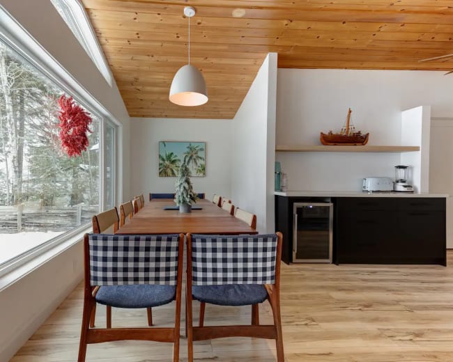 A long wooden dining table with checkered chairs is positioned next to large windows that overlook a snowy landscape, while a modern kitchen area is situated in the background.