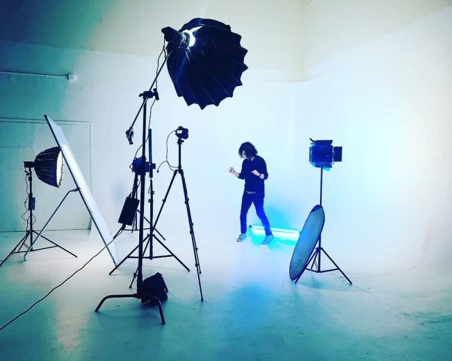 A person is dancing in a well-lit photo studio surrounded by various lighting equipment on a white backdrop.