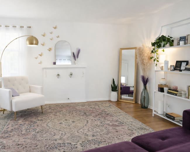 The room features a white-paneled wall with a decorative mirror, a plush white armchair, and a large area rug, complemented by plants and decorative elements on shelves.