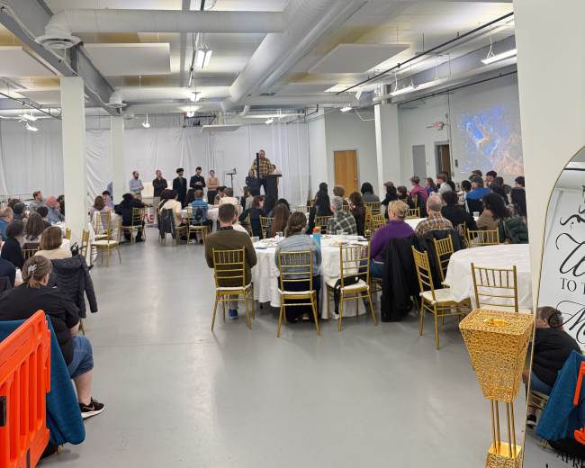 A large group of people sits at elegantly set tables in a spacious event hall with a stage at the front.