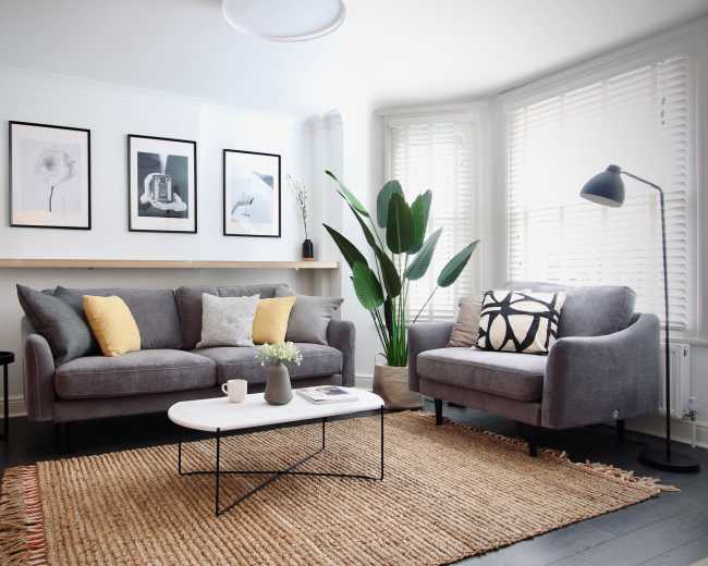 The image displays a modern living room with a gray sofa, white coffee table, and framed artwork on the walls, complemented by indoor plants and a natural fiber rug.