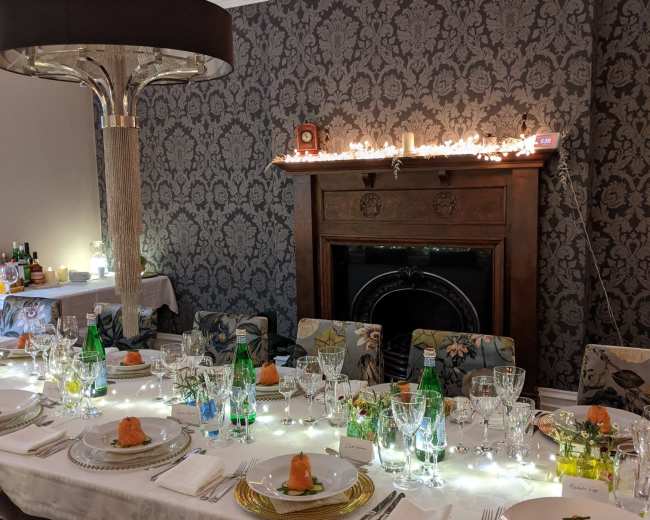 A long, elegantly set dining table with white tablecloth, gold-rimmed plates, and soft lighting in a patterned room.