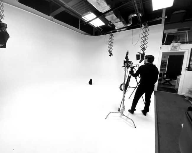 A person operates a camera in a spacious, well-lit studio with a white backdrop and equipment setup for a shoot.
