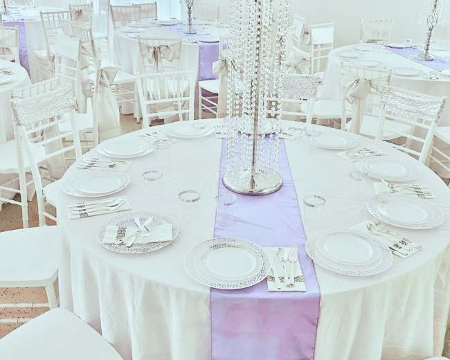 A finely decorated banquet hall features round tables set with white tablecloths, purple runners, and floral centerpieces, surrounded by white chairs.