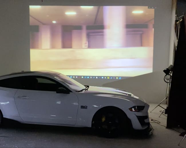 A white car is parked in a dimly lit room with a large projection of an indoor scene displayed on the wall behind it.