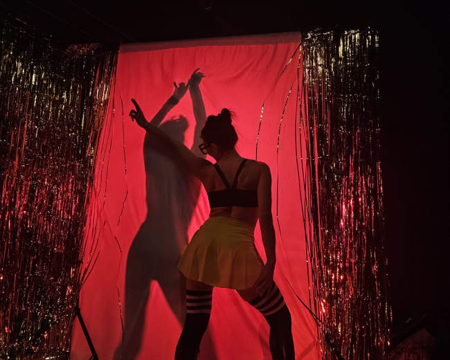 A silhouette of a person in a black top and a yellow skirt poses against a red-lit background with golden streamers.