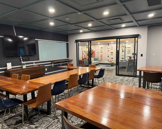 The image shows a modern conference room featuring several wooden tables and chairs, a large television, and glass doors leading to an area with colorful walls and furniture.
