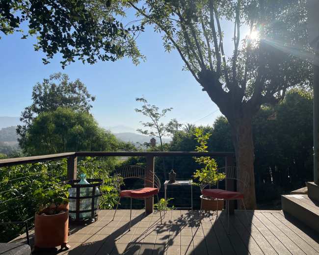 A wooden deck with two red chairs overlooks a green garden and distant hills under a clear blue sky.