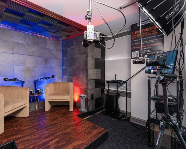 The image shows a video studio set up with two chairs, microphones, and camera equipment against a backdrop of textured gray walls and colored lighting.