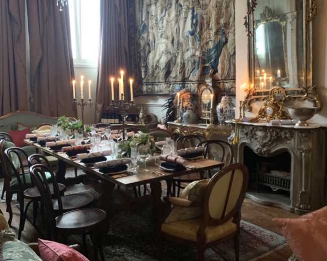 The picture shows an elegantly arranged dining room featuring a large wooden table set with dishes and candles, surrounded by various seating and decorated with a tapestry and a mirror.