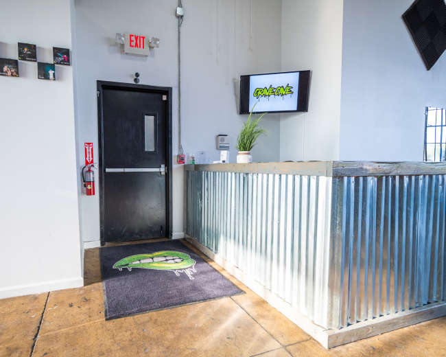 The entrance of a venue with a metal reception desk, a flat-screen TV on the wall, and a welcome mat featuring a graphic design.