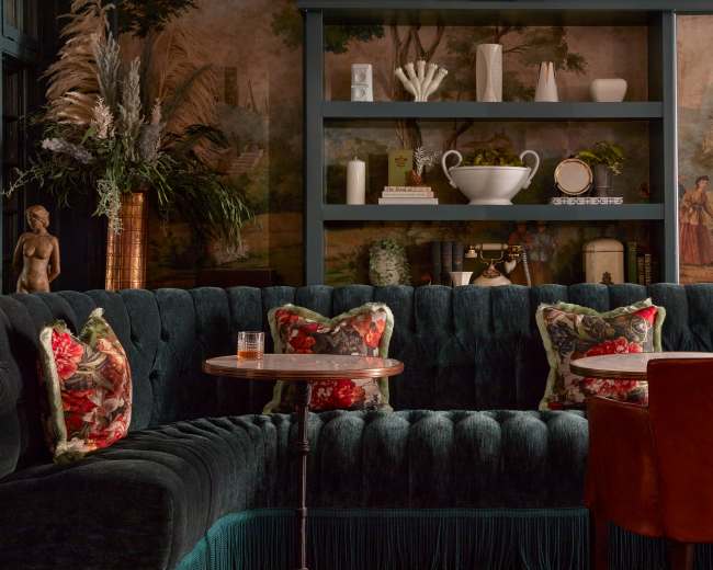 The image depicts a stylish lounge area featuring a plush green velvet sofa adorned with floral-patterned pillows, small round tables, and decorative shelves with plants and art.