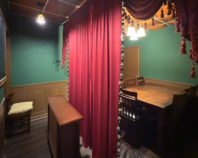 The image shows a dimly lit room with a wooden table surrounded by chairs, separated by a red velvet curtain.
