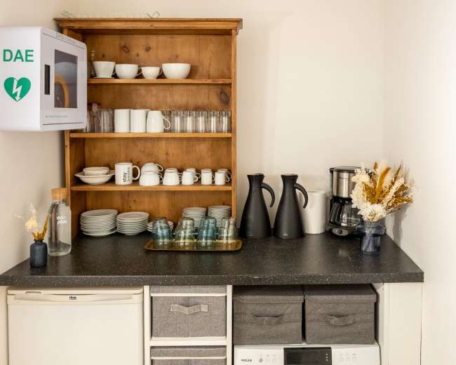 A minimalist kitchen corner features a refrigerator, a countertop with various dishes and glassware, and a stacked drawer unit beneath a microwave and coffee maker.